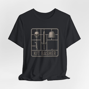 Może przedstawiać: Czarny t-shirt z nadrukiem graficznym przedstawiającym szarą wypraskę z plastikowymi częściami modelarskimi. Wypraska zawiera czaszkę, miecz, nóż i naramiennik. Pod częściami modelu znajduje się napis KIT BASHER wykonany szarą czcionką.