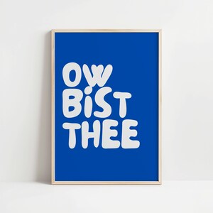 Può includere: Una stampa artistica incorniciata con uno sfondo blu vibrante. La stampa presenta il testo bianco "OW BIST THEE" in un carattere giocoso e arrotondato. L'opera d'arte è racchiusa in una semplice cornice di legno chiaro, adatta per l'arredamento murale.