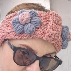 May include: A knitted headband in a soft pink hue, adorned with a large flower in shades of gray and pink. The headband features a textured knit pattern and is designed to be worn around the head.