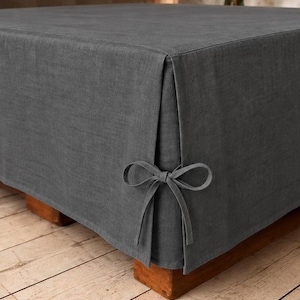 Può includere: Un copri-pouf in tessuto grigio scuro drappeggiato su un poggiapiedi quadrato, caratterizzato da una piega angolare sartoriale fissata con un laccetto in tessuto abbinato. Il tessuto ha una trama visibile e copre interamente il mobile.