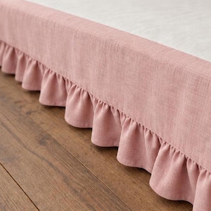Può includere: Una balza per letto color rosa antico caratterizzata da una finitura in tessuto testurizzato e da un bordo con volant arricciato. La balza è posizionata sotto un materasso bianco, creando un bordo decorativo che pende verso il pavimento.