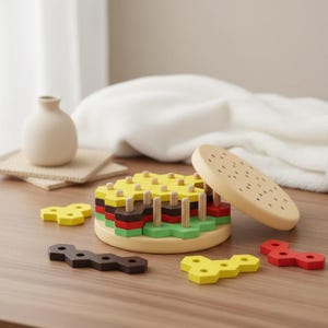 Puede incluir: Un juguete de madera para apilar con forma de hamburguesa, con capas de colores amarillo, rojo, marrón y verde. Los panes superior e inferior son de color marrón claro con pequeños agujeros. Varias piezas de madera sueltas están dispersas.