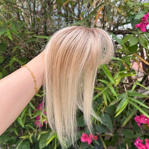 Könnte beinhalten: Ein blondes Haarteil mit einem hellbraunen Ansatz. Das Haarteil hat lange, glatte Strähnen und wird vor einem Hintergrund aus grünem Laub und rosa Blüten gehalten. Das Haarteil ist so konzipiert, dass es Volumen und Länge verleiht.