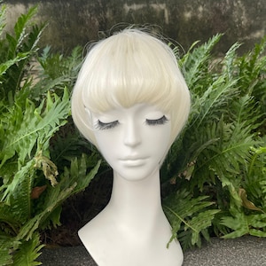 May include: A white mannequin head wearing a short, platinum blonde wig with straight bangs. The wig features a smooth, rounded bob style that frames the face, while the mannequin has closed eyes with long, dark, defined eyelashes.
