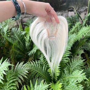 May include: A hair topper piece featuring straight, platinum white hair attached to a small, beige, mesh-like base. The base includes three silver-toned metal pressure-sensitive clips designed for secure attachment to natural hair.