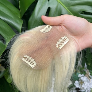 Op de afbeelding: Een platinablond haarstukje met een transparante, huidkleurige kanten basis en drie kleine metalen clips aan de onderzijde voor een stevige bevestiging. De steile, lichtgekleurde haarlokken lopen uit vanaf de basis.