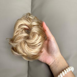 May include: A synthetic hair scrunchie in a light blonde shade with multi-tonal highlights. The hairpiece features a voluminous, wavy, and textured design arranged in a circular bun shape, providing a full appearance for styling hair.