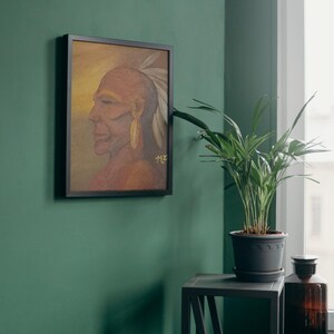 Puede incluir: Pintura enmarcada de un hombre nativo americano de perfil, con un tocado de plumas y un pendiente dorado. La obra de arte tiene un fondo cálido y dorado y está firmada. El marco negro cuelga de una pared verde.