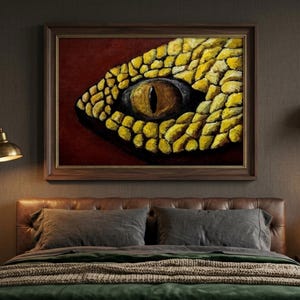Puede incluir: Un cuadro enmarcado que representa el ojo de una serpiente, con escamas amarillas y una pupila oscura y ovalada. La obra de arte está sobre un fondo rojo oscuro y enmarcada en madera oscura. El cuadro cuelga sobre una cama con almohadas grises y una manta verde.