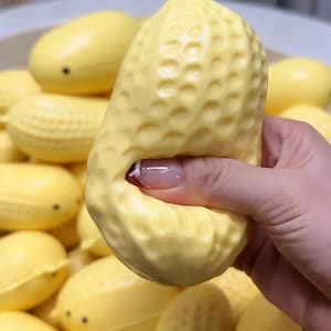 Puede incluir: Una bola antiestrés con forma de cacahuete amarillo claro con una superficie texturizada está siendo apretada por una mano. Otras bolas antiestrés similares están en el fondo. El objeto es divertido y táctil.