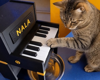 May include: A black and yellow pet feeder shaped like a miniature upright piano with the name NALA printed in yellow letters on the front panel. The piano features a set of black and white keys and a circular stainless steel bowl underneath.