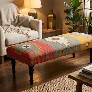 Puede incluir: Un banco rectangular tapizado con una superficie de tela tejida que presenta un patrón geométrico en tonos rojo, gris, amarillo y crema. El banco se apoya sobre cuatro patas de madera torneada de color marrón oscuro para mayor estabilidad.