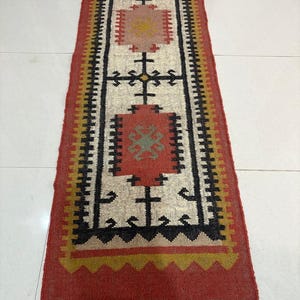 May include: A rectangular runner rug featuring a cream-colored center with geometric patterns in red, black, and yellow. The rug has a wide red border, a serrated yellow edge, and a fringe of grey tassels at the bottom end, with a rolled top section.