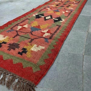 May include: A rectangular runner rug featuring a terracotta red base with a repeating geometric floral pattern in shades of olive green, mustard yellow, black, and cream. The rug has a jagged green border and brown fringe at the ends.