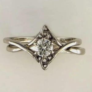 May include: A silver engagement ring with a round, clear gemstone at the center. The ring features a Celtic knot design around the gemstone and on the band. The ring is set against a white background.