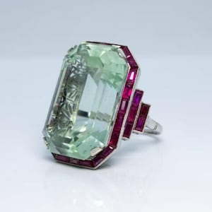 Pode incluir: Um anel com uma grande pedra verde retangular e uma banda prateada. A pedra é emoldurada por pequenas pedras vermelhas retangulares. O anel apresenta um design geométrico, com uma disposição escalonada de pedras vermelhas na lateral.