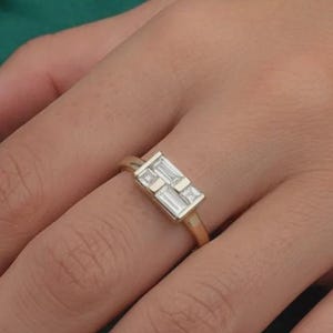 May include: A gold ring with two rectangular, clear gemstones. The ring is on a hand with pink nail polish, set against a dark green background.