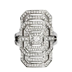 May include: A silver-toned ring with a rectangular, Art Deco-inspired design. The ring is encrusted with numerous small, clear gemstones, creating a sparkling effect. The central design is a series of geometric shapes, with a larger gemstone at the center.