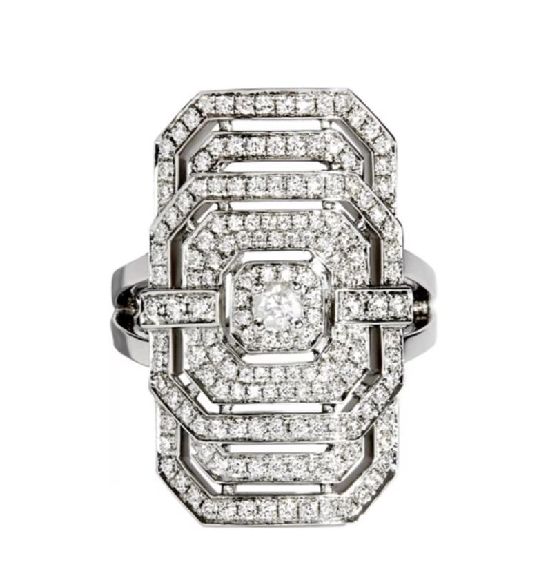 May include: A silver-toned ring with a rectangular, Art Deco-inspired design. The ring is encrusted with numerous small, clear gemstones, creating a sparkling effect. The central design is a series of geometric shapes, with a larger gemstone at the center.
