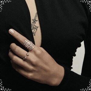 May include: A hand wearing three rings. The largest ring is rose gold with an intricate, layered design. Two smaller rings are also visible. The person is wearing a black long-sleeved top. A tattoo of a sword and flower is visible on the chest.