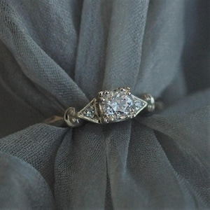 May include: A silver engagement ring with a large, round, clear diamond center stone. The ring has a vintage design with smaller accent diamonds on either side of the center stone. The ring is set against a soft, gray background.