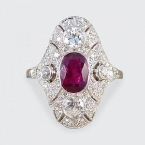 May include: An antique-style ring featuring a large oval ruby center stone, surrounded by a halo of small diamonds. Two larger diamonds are set above and below the ruby, with additional diamond accents on the ring's shoulders. The ring has a silver-colored band.
