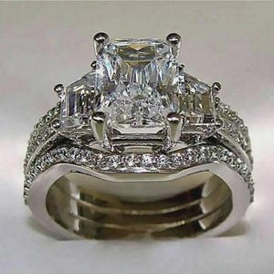 May include: A dazzling engagement ring set with a large, rectangular-cut center stone, flanked by two smaller stones. The band is encrusted with numerous small, sparkling accent stones, creating a luxurious and eye-catching design. The ring is made of a shiny, silver-colored metal.