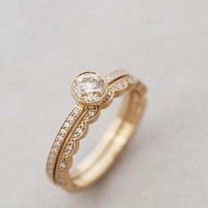 May include: A gold engagement ring set with a round, clear gemstone and a matching band. The engagement ring has a bezel setting and is surrounded by small, sparkling stones. The band has a scalloped edge and is also adorned with small stones.