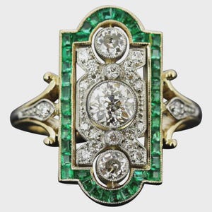 May include: An Art Deco ring with a gold band, emeralds, and diamonds. The rectangular design features three large diamonds in the centre, surrounded by smaller diamonds. Rectangular emeralds frame the diamonds, creating a striking contrast. A vintage-inspired piece.