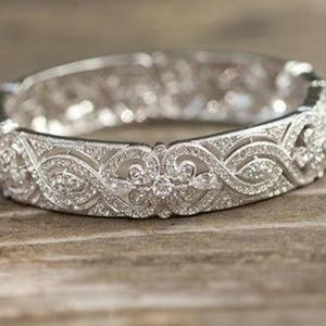 Può includere: Un braccialetto rigido argentato con intricati filigrane e abbellimenti scintillanti. Il braccialetto presenta un motivo ripetuto di disegni a spirale e accenti floreali, creando un aspetto lussuoso ed elegante. Il braccialetto è appoggiato su una superficie di legno.