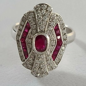 Pode incluir: Anel de prata com um rubi oval central, rodeado por pequenos diamantes e rubis retangulares. O anel apresenta um design Art Déco com uma forma geométrica e uma banda polida.