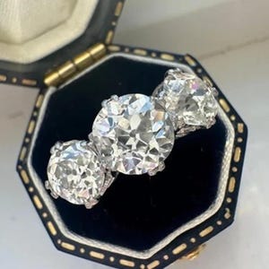 Puede incluir: Un anillo de diamantes de tres piedras con una piedra central grande y redonda y dos piedras laterales más pequeñas. El anillo está engastado en una banda plateada y se muestra en un joyero negro y dorado. Los diamantes brillan.