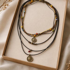 Op de afbeelding: Een meerstrengs ketting met donkerbruine en zwarte koorden, geaccentueerd met kleine, kleurrijke glaskralen in geel, rood, blauw en groen. Het sieraad bevat twee ronde, antiek-bronskleurige metalen hangers met gedetailleerde reliëfpatronen.
