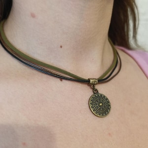 Puede incluir: Un collar de varias hebras con tres cordones finos en verde oliva, negro y marrón. El collar incluye un colgante circular de metal en tono bronce antiguo con un patrón geométrico de rayos de sol y una anilla de metal texturizada a juego.