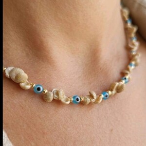 Puede incluir: Un collar con conchas marinas naturales, pequeñas cuentas doradas y cuentas azules en forma de ojo. El collar está diseñado para ser usado alrededor del cuello. Las conchas son beige y las cuentas de ojo son de un azul brillante.