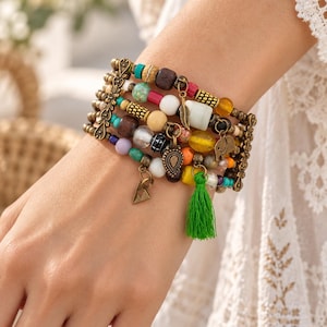 Puede incluir: Una pulsera de múltiples hilos con un surtido de cuentas coloridas en tonos turquesa, amarillo, marrón, blanco y naranja. La pieza incluye espaciadores de metal en tono bronce antiguo, dijes con forma de pluma, elefante y triángulo, y una borla de color verde brillante.