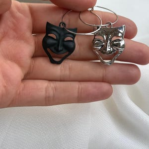 Può includere: Un paio di orecchini a cerchio con ciondoli a forma di maschera teatrale. Un ciondolo è nero opaco e l'altro è argento lucido. Le maschere raffigurano un'espressione sorridente con dettagli su occhi, naso e bocca, appese a sottili cerchi metallici.