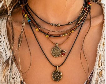 May include: A multi-layered necklace featuring black leather cords, thin metallic chains, and small colorful beads. The piece includes two bronze-toned circular pendants with intricate embossed patterns and small dangling red and gold accents.