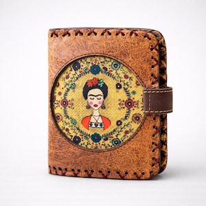 Puede incluir: Cartera de cuero marrón con un retrato de una mujer con una corona de flores y una blusa roja. La cartera tiene un borde cosido y un cierre de correa de cuero marrón. El retrato está rodeado de un diseño floral.