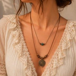 May include: Two layered necklaces. The top necklace has a string of small turquoise beads. The bottom necklace has a circular pendant with an intricate design and wooden beads. The necklaces are worn over a white blouse with lace trim.