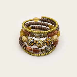 May include: A multi-strand wrap bracelet featuring a combination of brown wooden disc beads, olive green seed beads, and three central focal beads with a black, white, and yellow speckled pattern, accented by metallic gold-toned spacers.