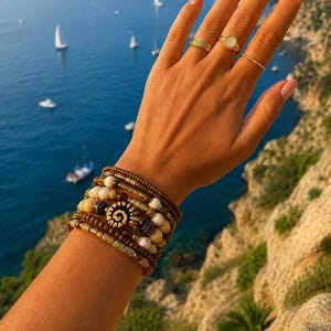 Puede incluir: Una mano con anillos dorados y pulseras de cuentas apiladas. Las pulseras presentan una variedad de cuentas marrones, beige y blancas, con un diseño central en espiral. El fondo muestra un mar azul con veleros y un acantilado rocoso.