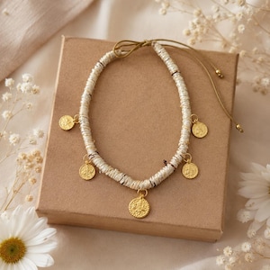 Puede incluir: Un collar que presenta una hilera de pequeñas cuentas cilíndricas de color blanco roto con cinco dijes circulares dorados en forma de moneda que cuelgan a intervalos. El collar incluye un cierre de cordón marrón con cuentas doradas.