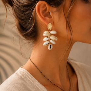 Puede incluir: Pendientes dorados con un perno ovalado texturizado y una cascada de acentos de concha blanca y crema. También se ve un delicado collar de oro con un colgante. Los pendientes son un accesorio único.