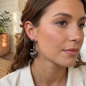 May include: A dark metal earring featuring a star, a sword, and a heart-shaped pendant. The heart has a decorative design and a red gem. The earring is a dangle style.
