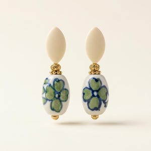 Puede incluir: Un par de pendientes colgantes con una parte superior ovalada color crema, detalles dorados y una cuenta ovalada blanca con un diseño floral azul y verde. Un accesorio único.