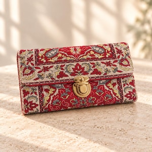May include: A rectangular wallet featuring a woven fabric exterior with a red, beige, and grey floral pattern. The wallet is secured with a polished gold-tone metal push-lock clasp centered on the front flap, showcasing intricate textile details.