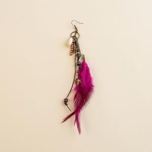 May include: A single, handcrafted earring featuring a vibrant fuchsia feather. The earring includes a metal hook, beads, a small charm, and a peace sign pendant. The design has a bohemian aesthetic.