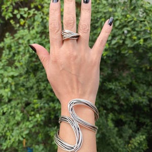 Könnte beinhalten: Ein silberfarbenes, mehrsträngiges Armband mit einer Reihe überlappender, kreisförmiger Drahtschleifen und ein passender mehrsträngiger Ring mit gekreuztem Design, beide gefertigt aus dünnen, polierten Metalldrahtelementen.