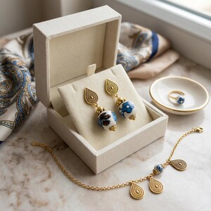 Puede incluir: Un conjunto de joyas con pendientes, pulsera y anillo dorados. Los pendientes y la pulsera tienen cuentas florales azules y blancas. El anillo está colocado en un pequeño cuenco blanco con borde dorado. Los pendientes se muestran en una caja color crema.